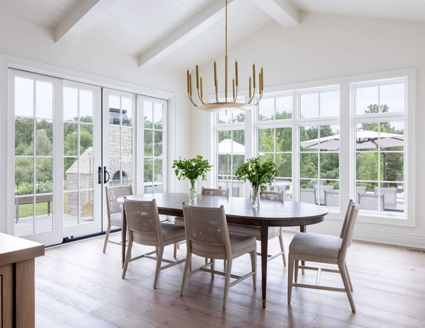 Bright, spacious dining room with a large dark wood dining table and upholstered dining chairs, surrounded by extensive white-framed windows and glazed French doors. The room has white walls and a white vaulted/beamed ceiling, pale timber flooring, and a sculptural brass-gold linear chandelier. Outdoor patio and greenery are visible beyond the glazing.
