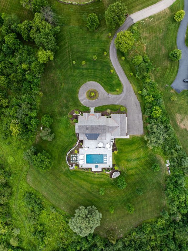 A vertical aerial, top-down view of a large landscaped residential property. The house has a grey pitched roof and a rear terrace with outdoor furniture surrounding a rectangular swimming pool. Curving paved driveways, a circular turning area, extensive manicured lawns, mature trees, and dense woodland frame the site.
