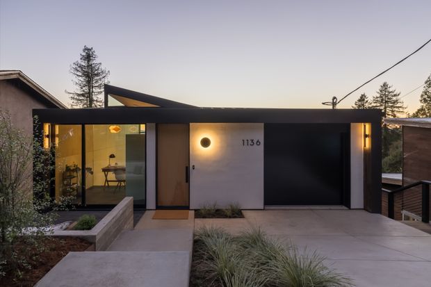 Twilight exterior view of a compact contemporary residence with a strong horizontal dark façade, flat roofline, warm recessed and wall lighting, and a glazed entry volume. A concrete walkway leads through low landscaped planting to the front door, while the adjacent garage or large dark opening is integrated into the façade. The house sits among mature trees and neighbouring buildings on a slightly elevated site.