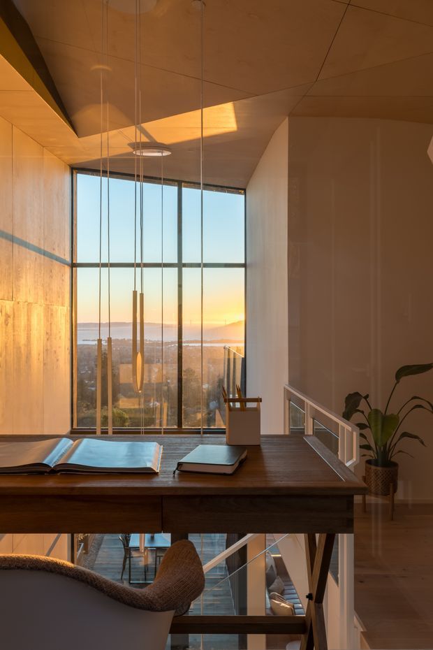 A warm, contemporary interior view of a home office or study positioned beside a double-height glazed stairwell. A dark timber desk with books occupies the foreground, while full-height windows reveal a sunlit coastal or waterfront outlook. Slim pendant lights hang from a timber-paneled ceiling, and a stair with a simple light balustrade descends to a lower level. The space uses strong vertical glazing, warm wood tones, and sunset light to create an elegant, calm atmosphere.