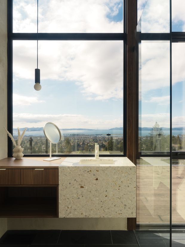 A refined bathroom vanity set against expansive floor-to-ceiling glazing, with a dark timber floating cabinet, pale terrazzo or stone benchtop and integrated basin. A round mirror and pendant light sit in front of the window, which frames a broad coastal or lakeside landscape. The space has strong natural light, dark tiled flooring, timber framing and a clean contemporary feel.