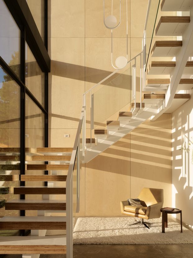 A sunlit contemporary stairway and double-height landing with floating timber stair treads, slim white stringers, clear glass balustrades, and warm pale timber wall panelling. Large black-framed glazing runs along the left side, while exposed timber beams and additional stair flights are visible above. A tan lounge chair, small dark side table, and textured cream rug create a quiet seating nook beneath the stairs. Strong diagonal shadows and pendant globe lights add visual interest.