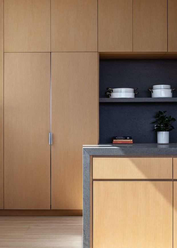 A close-up view of a refined, minimalist kitchen featuring extensive light timber cabinetry, a dark charcoal open shelving and splashback zone, and a dark grey stone benchtop. White cookware, a small potted plant, and a book sit on the open shelves. Warm sunlight creates strong diagonal highlights across the cabinetry and floor, emphasizing the clean built-in joinery and restrained material palette.