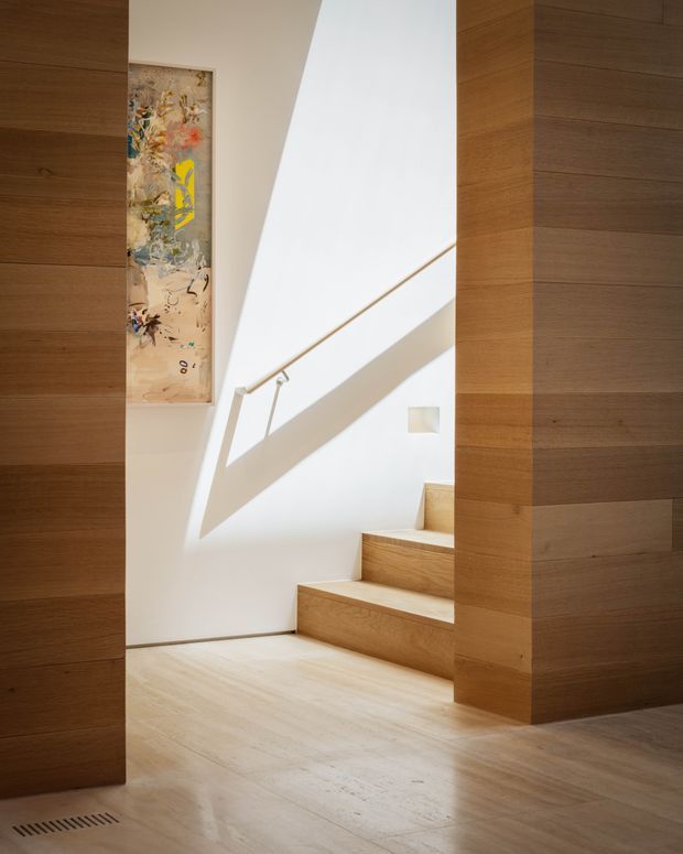 A bright minimalist interior stairway with broad timber flooring, timber-clad walls and stair treads, white walls, and a slim metal handrail. Strong daylight enters from an upper opening or skylight, casting geometric shadows across the stairwell. An abstract artwork is mounted on the wall beside the stairs.