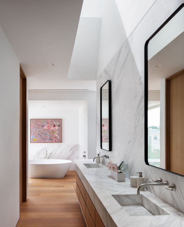 A refined contemporary bathroom with a long marble vanity, twin integrated stone basins, wall-mounted brushed metal taps, and large rounded-corner black-framed mirrors. Warm timber cabinetry and timber flooring contrast with the pale veined stone surfaces. Beyond the vanity is a freestanding white bathtub set against a marble feature wall, with a pink artwork above it. The space is bright, minimal, and luxurious, with a high ceiling and strong vertical architectural volume.