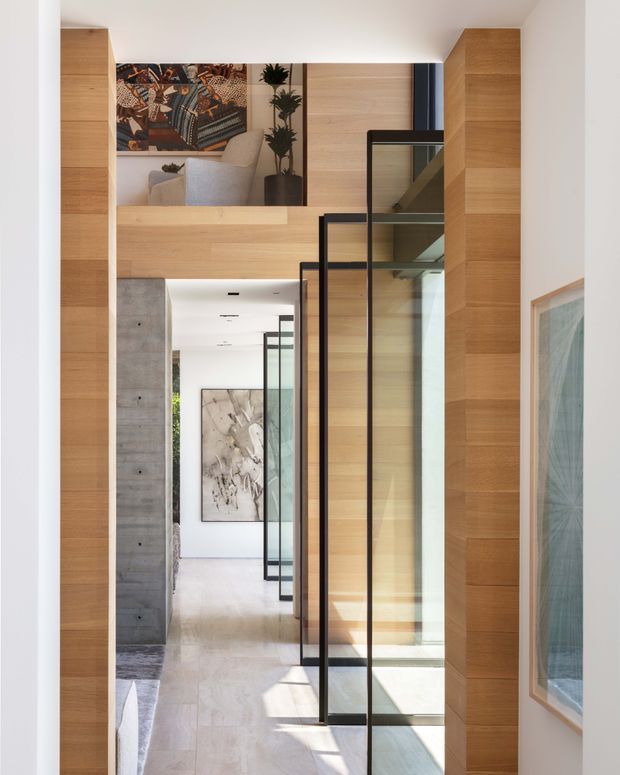 A bright, refined interior view through a tall hallway or entry passage. Warm light-toned timber wall panelling and built-in joinery frame a sequence of slim black-framed glass partitions and doors. A concrete wall, pale stone or concrete flooring, artwork, a partial lounge area, and a glimpse of a staircase contribute to the contemporary architectural composition.
