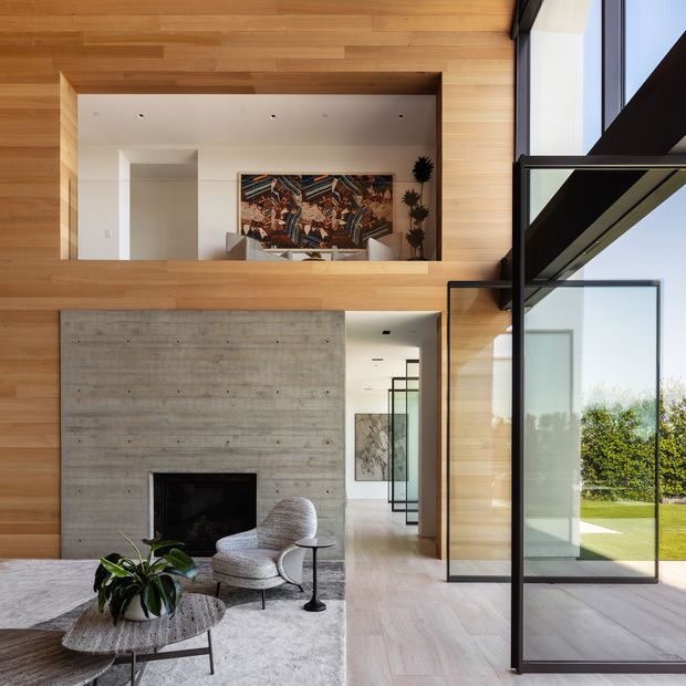 A refined double-height living room with warm timber wall and ceiling panelling, a large exposed concrete fireplace, and full-height black-framed glazing opening toward a lawn. A mezzanine-like upper sitting area is visible above the fireplace, with contemporary lounge furniture, a sculptural rug, artwork, and indoor plants. The space has strong natural light and a seamless indoor-outdoor connection.