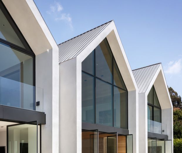 Exterior architectural photograph of a contemporary multi-volume building with repeated steep gabled forms, crisp white rendered walls, dark metal-framed floor-to-ceiling glazing, and standing-seam metal roofing. The facade is viewed from a low angle against a blue sky, with transparent entry and living areas visible through the glazing.