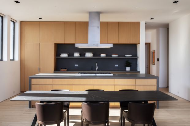 A contemporary open-plan kitchen and dining area with extensive light timber cabinetry, a dark charcoal splashback, and a large dark stone island bench. A long dark dining table with brown upholstered chairs occupies the foreground. The kitchen has integrated appliances, a central stainless-steel rangehood, under-cabinet lighting, pale timber flooring, recessed ceiling lights, and bright windows along the left wall.