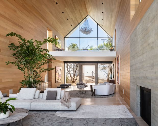 A spacious double-height living room with a dramatic glazed gable at the upper level, warm timber-clad walls and ceiling, a large concrete or stone fireplace wall, and an open mezzanine balcony. The room contains a pale sectional sofa, freestanding bathtub-like sculptural seating or tub near the fireplace, contemporary lounge furniture, indoor trees, artwork, and a large neutral rug. Abundant glazing brings in daylight and overlooks palms and landscaping.