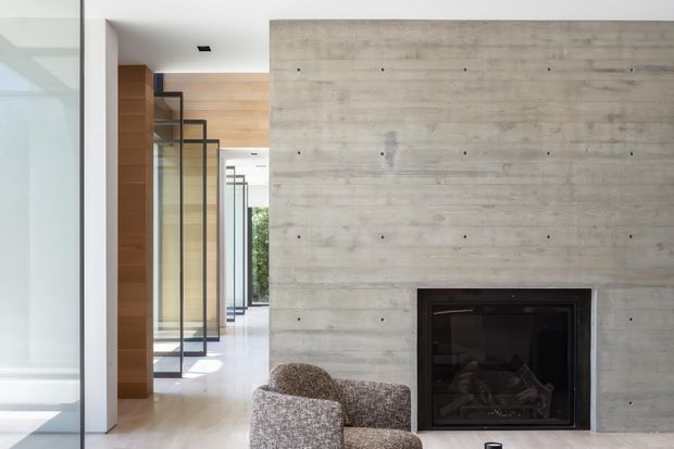 Contemporary interior focused on a broad pale concrete feature wall with an integrated dark fireplace. A pair of textured lounge chairs sits in the foreground, while a bright corridor beyond features timber wall panelling and repeated black-framed glazed doors or partitions. The space has clean linear detailing, polished light flooring, and abundant natural light.