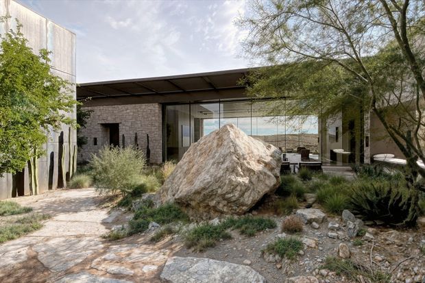 Exterior view of a low-slung modern residence integrated into a rocky, landscaped site. The building features dark stone masonry walls, extensive floor-to-ceiling glazing, a broad flat roof canopy, and a large natural boulder prominently positioned in the foreground. Native-looking drought-tolerant planting, stone paving, and mature trees surround the house.