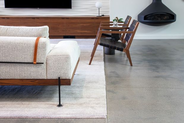 A refined mid-century-inspired living room featuring a low upholstered sofa, built-in timber media cabinetry, a timber lounge chair with integrated shelving, a suspended black fireplace, and a polished concrete floor. The room has a restrained neutral palette with warm wood accents and a large textured rug.