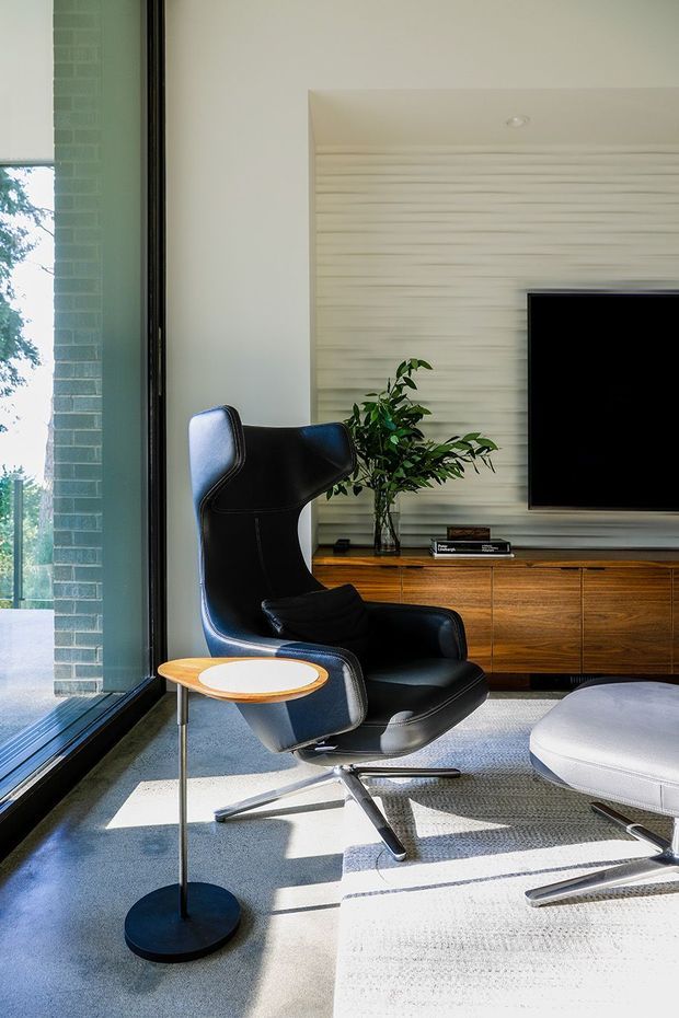A bright, refined living room featuring a black leather lounge chair with matching ottoman, a slim round side table, a timber media console, wall-mounted television, and a horizontally textured feature wall. Full-height glazing at the left brings in strong natural light and views of greenery. The room has a soft grey carpet and a restrained mid-century-inspired furniture arrangement.