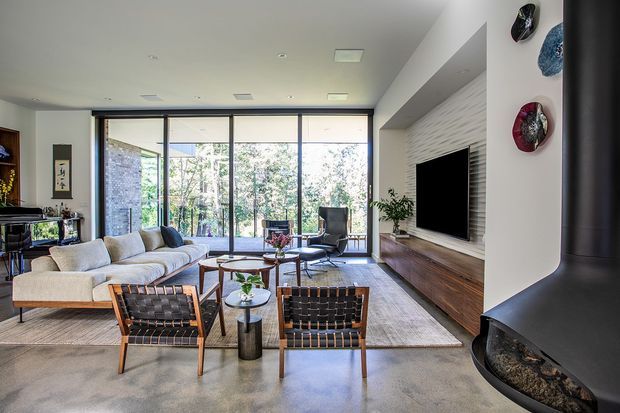 Bright contemporary living room with a large beige sectional sofa, leather-and-wood lounge chairs, a central glass coffee table, and a wall-mounted television integrated into a long timber media console. Full-height glazed doors open the room to a leafy outdoor setting. A sculptural black suspended fireplace occupies the right foreground, while textured white wall cladding and decorative wall objects add visual interest.