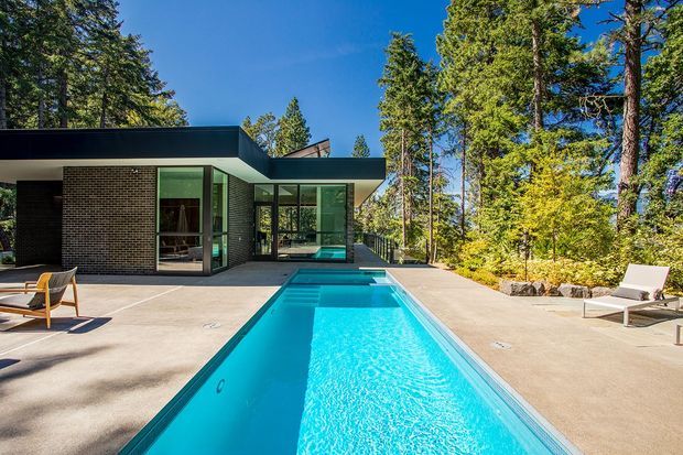 A modern, single-storey flat-roof house set within a wooded landscape, viewed from a spacious paved terrace. The foreground is dominated by a long rectangular swimming pool, with full-height glazing, dark brick walls, and broad roof overhangs forming the house facade. Outdoor lounge chairs sit beside the pool, and mature trees surround the property.