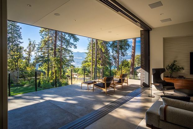 A contemporary living room opens onto a generous outdoor terrace through fully retractable, floor-to-ceiling glazed doors. The room features a neutral carpeted seating area, a dark sculptural lounge chair, a sofa, timber built-in cabinetry, and a large television. The covered terrace has concrete paving, glass balustrades, and timber-framed outdoor lounge chairs, with a wooded landscape and distant hills visible beyond.