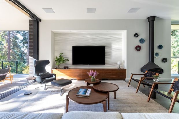 Bright, mid-century-inspired living room with expansive glazing to a wooded exterior. The room features a large textured white media-wall niche with a television, low timber sideboard, sculptural lounge chair and ottoman, nesting round timber coffee tables, neutral rug and sofa, and a black freestanding fireplace with a tall flue. Decorative wall plates and indoor plants add restrained colour.