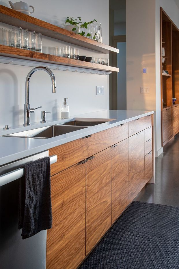 A contemporary kitchen with a long white countertop, double stainless-steel sink and chrome gooseneck tap. Warm natural timber cabinetry with visible grain and dark pulls runs beneath the bench, while open timber shelves display glassware, crockery and plants. The space has white walls, dark flooring and a partially visible timber-lined adjoining area.