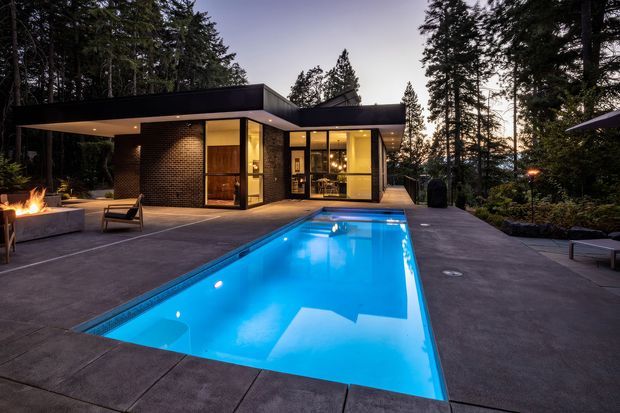 A modern single-storey pavilion-style house set among tall trees, photographed at dusk. The flat-roofed building has extensive floor-to-ceiling glazing, dark brick walls, warm interior lighting, and a broad concrete terrace surrounding a long illuminated swimming pool. An outdoor fire feature and lounge seating create a dramatic, resort-like outdoor setting.