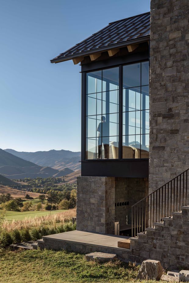 Exterior architectural photograph of a stone-clad mountain home featuring a projecting glazed room with large black-framed grid windows, exposed timber eaves, and a dark pitched tiled roof. A stone stair and paved terrace sit beside the building, with a broad valley and mountains visible beyond.