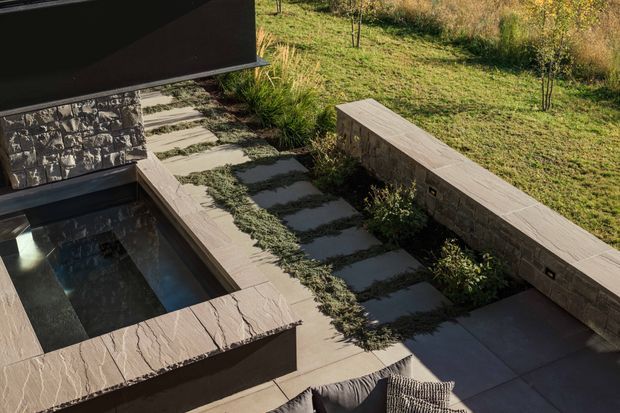 A landscaped outdoor courtyard beside a modern building, viewed from above. The scene features a compact dark-lined reflecting or plunge pool, broad natural stone coping, a stepped pathway of large dark stone slabs set among groundcover, and a low stone masonry wall with a smooth stone cap. Grasses and understated planting surround the hardscaped area, creating a restrained contemporary garden.