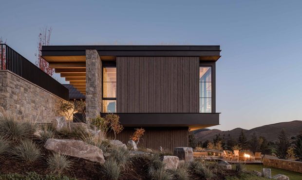 Exterior architectural photograph of a contemporary multi-level residence set into a sloped, rocky landscape. The house features dark vertical timber cladding, large glazed openings, strong horizontal roof planes, exposed warm timber soffits, and substantial natural stone walls and columns. Terraced outdoor areas and rugged hills are visible around the building at dusk.