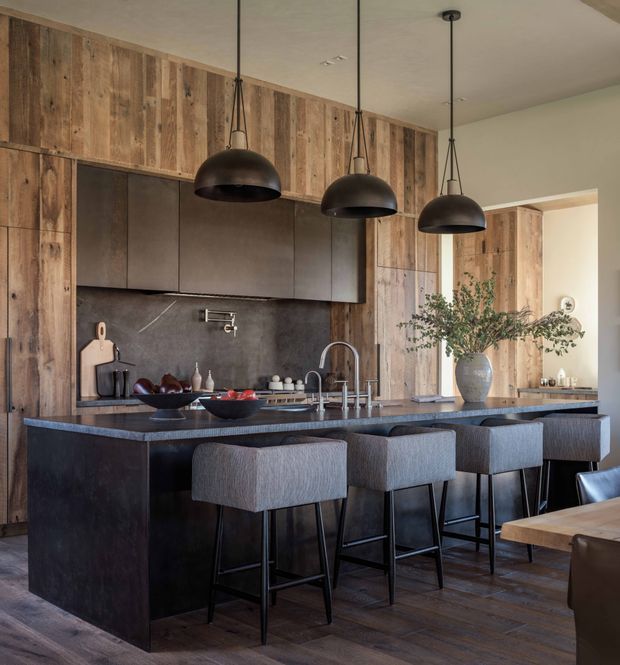 A sophisticated open-plan kitchen dominated by a large dark island with waterfall-style sides and four upholstered bar stools. Full-height rustic timber wall panelling and matching timber cabinetry frame the kitchen, while dark matte cabinetry and a dark stone-look splashback provide contrast. Three oversized black dome pendant lights hang above the island. A dining area is partially visible at the right, with warm natural light and understated styling.