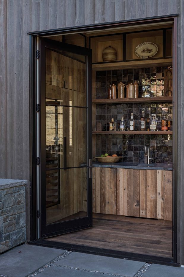 A compact, dark-toned built-in bar and storage area viewed from outside through an open black-framed glass door. The space features vertically grained timber wall cladding, timber cabinetry, open timber shelves, a dark glossy tiled splashback, a stone benchtop, integrated sink and tapware, bottles and ceramic vessels. The bar opens directly onto a paved exterior area and is framed by dark timber cladding and masonry.