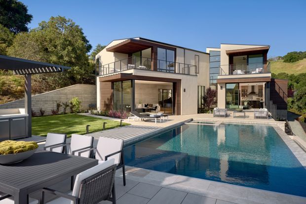 A contemporary multi-level residential home viewed from the landscaped pool area. The house has a rectilinear form with flat roof planes, light rendered walls, dark timber-toned soffits and trim, extensive floor-to-ceiling glazing, and multiple projecting balconies with slim dark metal balustrades. A large swimming pool dominates the foreground, with paved terraces and outdoor dining furniture. The property sits against a leafy, sloping suburban landscape.
