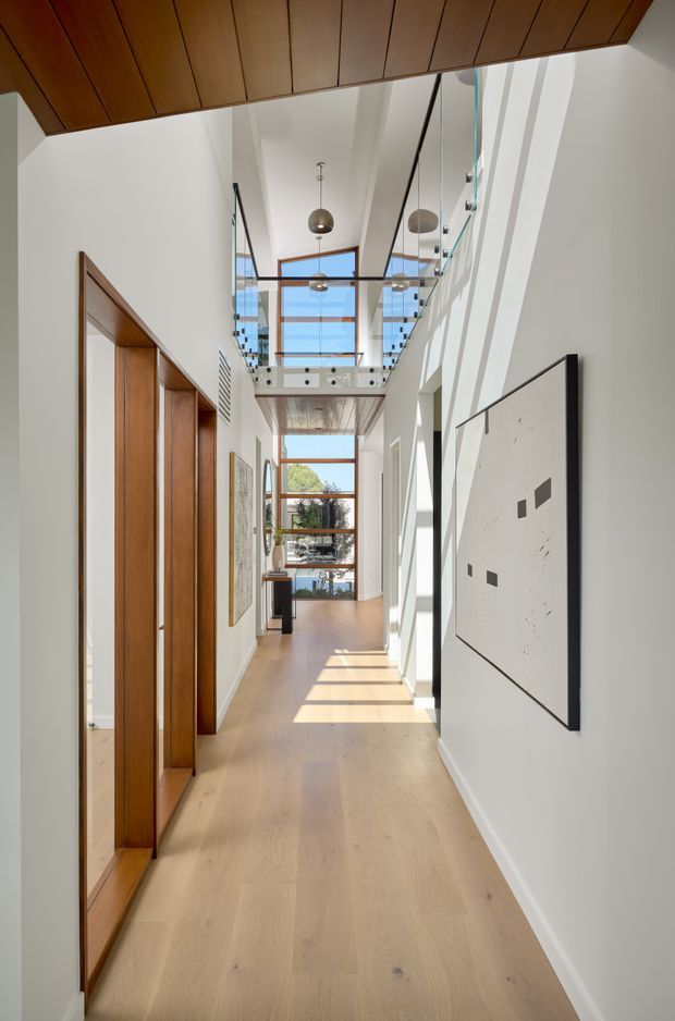 A bright, elongated residential hallway with pale timber flooring, white walls, tall timber-framed door and storage panels on the left, and a large minimalist artwork on the right. A glazed mezzanine or upper-level bridge with glass balustrades spans the corridor, creating a dramatic double-height void. Timber ceiling panelling, pendant lights, clerestory glazing, and full-height windows bring warmth and abundant natural light into the space.