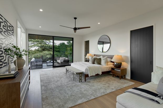 Bright contemporary bedroom with a low-profile upholstered bed, layered neutral bedding, timber bedside table, large area rug, lounge chair, and streamlined timber media cabinet. Floor-to-ceiling sliding glass doors open to a furnished balcony or terrace with greenery beyond. A black ceiling fan, recessed lighting, circular wall mirror, abstract wall art, and dark flush door add modern accents.