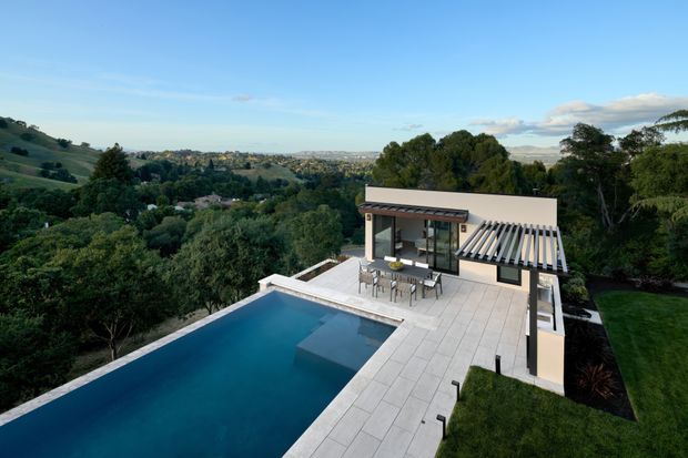 A contemporary hillside residence viewed from above, featuring a long rectangular swimming pool, expansive pale tiled terrace, and a compact pavilion with dark-framed glazing and a distinctive slatted roof canopy. The property is surrounded by mature trees and landscaped lawn, with broad suburban and rolling-hills views beyond. The composition is clean and geometric, with strong indoor-outdoor connection and a bright natural setting.