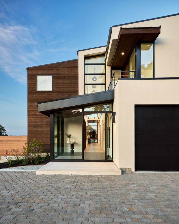 Exterior photograph of a contemporary multi-level residence with a flat-roofed composition, dark timber cladding, smooth light rendered walls, black-framed glazing, and a projecting timber-lined entry canopy. A glazed entrance reveals a bright interior hallway, while a large dark garage door and extensive grey paved driveway occupy the foreground.