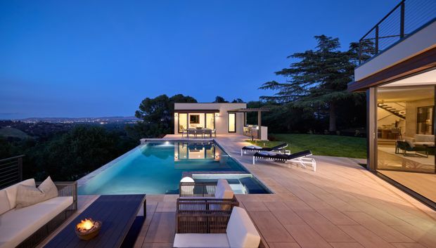 Twilight view of a luxurious modern home with a large rectangular swimming pool, expansive timber-toned outdoor decking, outdoor lounge furniture and fire bowl, opening to a glazed interior living area. The property sits on an elevated landscaped site with broad distant suburban views, while a small pavilion and poolside seating occupy the background.