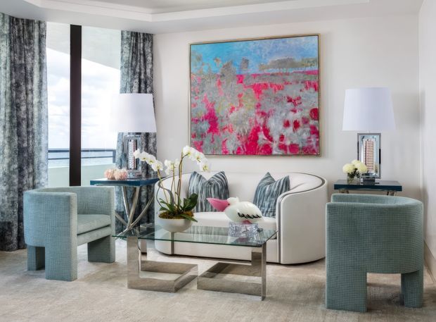 A refined contemporary living room with pale upholstered armchairs, a curved white sofa, glass and metal coffee table, large abstract artwork, sculptural lamps, orchids, and full-height glazing overlooking water. The room uses soft neutral finishes with blue-grey and vivid pink accents, creating an elegant, bright coastal feel.