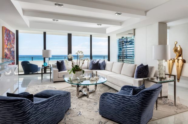 Bright, spacious contemporary living room with a large white sectional sofa, two navy patterned lounge chairs, glass coffee table, marble-toned flooring, decorative artwork, and sculptural accessories. Floor-to-ceiling glazing opens to an expansive ocean view, while recessed tray ceiling details and dark vertical window mullions add architectural definition.