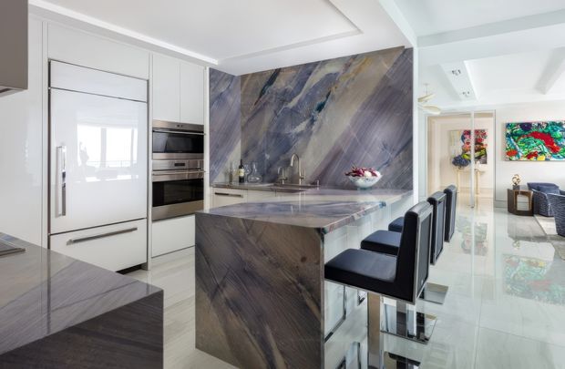 Bright, spacious contemporary kitchen with a large veined stone island and matching full-height splashback. White integrated cabinetry includes a tall refrigerator and built-in ovens, while dark upholstered bar stools line the island. The kitchen opens toward a light-filled living area with glossy flooring and colourful artwork visible beyond.