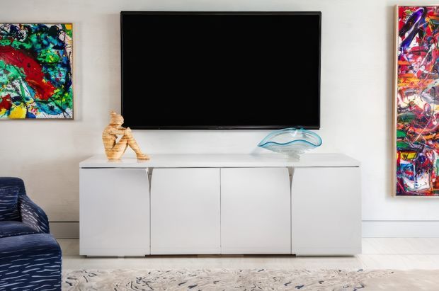 A contemporary living room media wall with a large wall-mounted black television above a sleek white sideboard. Abstract colourful artwork hangs on both sides, with sculptural decorative objects on the console, a patterned blue armchair at the edge of the frame, and a pale patterned rug on the floor.