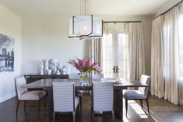 A bright, refined dining room with a dark timber dining table surrounded by upholstered, patterned dining chairs. Floor-to-ceiling cream curtains frame tall glazed doors and windows, while a contemporary geometric pendant light hangs above the table. A dark console with sculptural white decor and a floral arrangement completes the room.