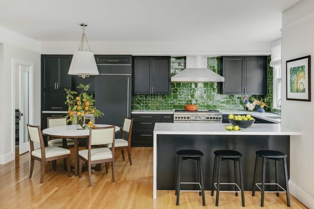 Bright open-plan kitchen and dining area with dark charcoal cabinetry, a large white island with three stools, stainless-steel appliances and rangehood, and a vivid green patterned tile splashback. A round dining table with upholstered wooden chairs sits beside the kitchen, with timber flooring and white walls creating a clean, contemporary feel.