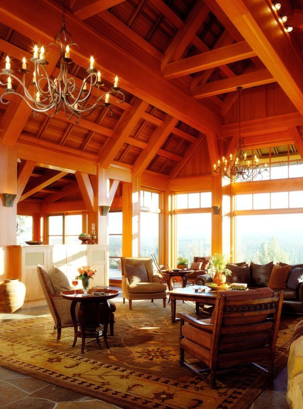 A spacious, warmly lit living room with dramatic exposed timber roof trusses, timber wall panelling, large windows, and multiple ornate chandeliers. The room is furnished with traditional woven and upholstered armchairs, a substantial carved timber sofa, dark wood tables, and patterned area rugs. Sunlight enters through the expansive glazing, with a distant landscape visible beyond.