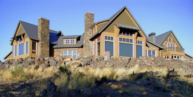 Exterior view of a large multi-gabled mountain-style residence on a rocky, elevated site. The house features steep pitched roofs with dark metal roofing, warm earth-toned wall cladding, prominent natural stone chimney towers, extensive multi-pane glazing, and timber-framed gable details.