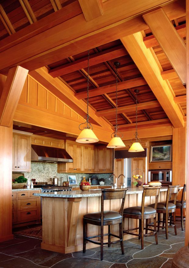 A warm, spacious timber-lined kitchen with a large central island and granite benchtop, integrated cooktop, extensive wood cabinetry, and a raised breakfast bar with four leather bar stools. The room features exposed timber beams and rafters, pendant lights, stone-patterned flooring, a stone mosaic backsplash, and tall timber-framed glazing and cabinetry. The high, heavily articulated ceiling and exposed structural framing give the space a lodge-like, rustic character.