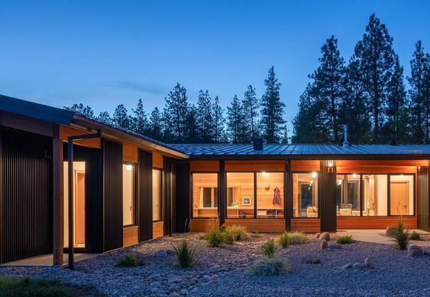 Twilight exterior view of a low-profile, single-storey contemporary home arranged around a landscaped gravel courtyard. The dark vertical-clad wings and broad metal roof frame warmly lit timber-toned interiors, with extensive glazing, timber posts, and sheltered outdoor circulation visible around the courtyard.