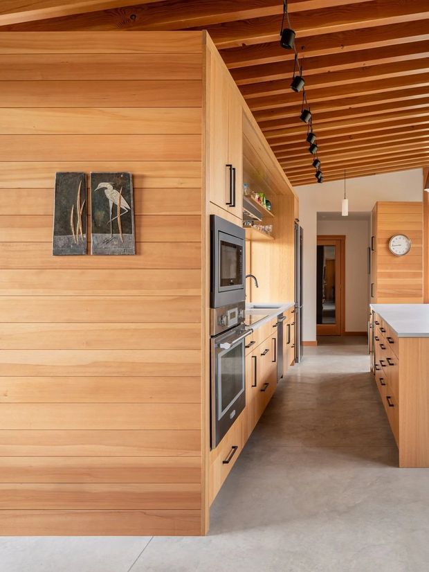 A warm, contemporary kitchen with extensive horizontal timber wall panelling and exposed timber ceiling battens. Built-in cabinetry runs along both sides of a galley-style layout, with integrated black ovens, refrigerator, sink, shelving, and pale benchtops. A central timber partition conceals appliances and storage, while black track-style pendant spotlights run along the ceiling. The space opens toward a hallway at the rear and has a polished concrete floor.