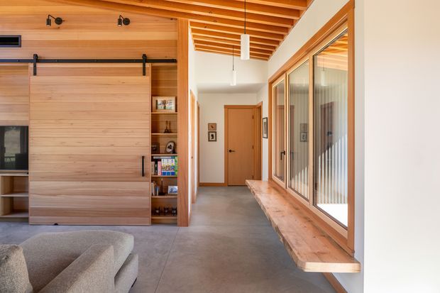 A warm, modern hallway and living area with extensive built-in timber joinery. The image shows a large sliding timber-panel partition on black hardware, open shelving, a partial sofa, a polished concrete floor, exposed timber ceiling battens, and a long timber window seat beside full-height glazed doors or windows. The corridor leads to a timber interior door, with strong natural light entering from the glazing.