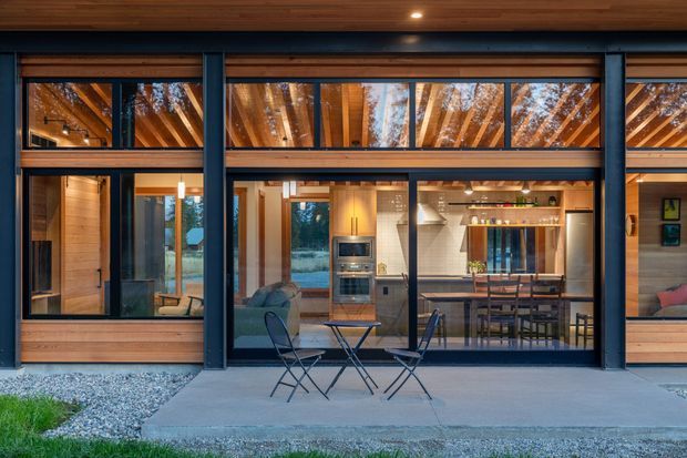A contemporary single-storey home viewed from an exterior patio, with extensive dark-framed glazing revealing a warm timber-lined interior. The central view shows an open-plan kitchen and living area, with a dining counter, pendant lighting, timber cabinetry, and exposed timber ceiling structure. A covered outdoor seating area with folding chairs occupies the foreground, while gravel landscaping borders the concrete patio.