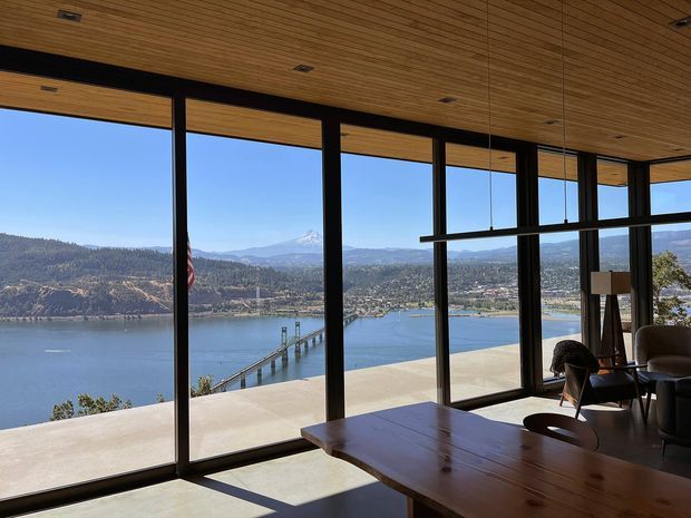 A bright contemporary interior with extensive floor-to-ceiling glazing overlooking a broad lake, shoreline settlement, wooded hills, and distant mountains. The room has a warm timber slatted ceiling, dark slender window and door frames, polished concrete flooring, and a large timber dining table with lounge seating. The dramatic waterfront outlook and clean structural glazing dominate the scene.