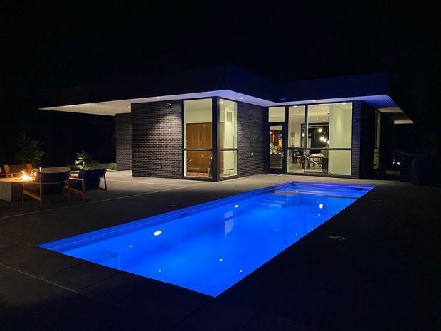 Night-time exterior view of a low-slung modern home with dark brick walls, extensive floor-to-ceiling glazing, and broad flat roof planes. A vividly illuminated rectangular swimming pool dominates the foreground, with a paved terrace, outdoor seating, and warm exterior lighting beside the house.